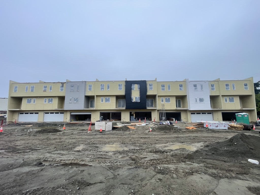 A row of townhomes under construction, showing framing, siding work, and the muddy ground, illustrating the development phase of the project.