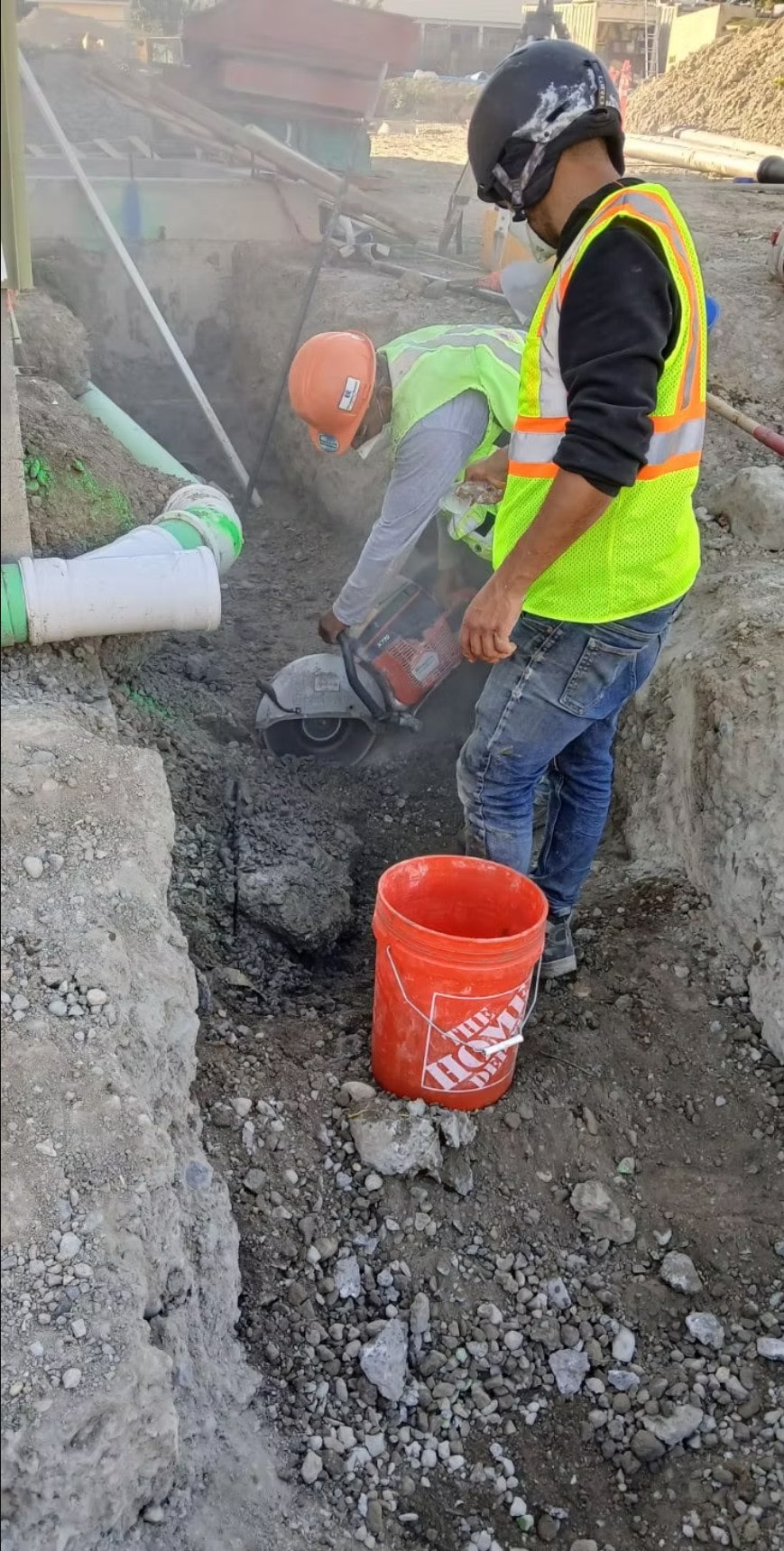 A construction worker uses a circular saw to cut concrete or pipe in a trench, highlighting specialized utility installation work.