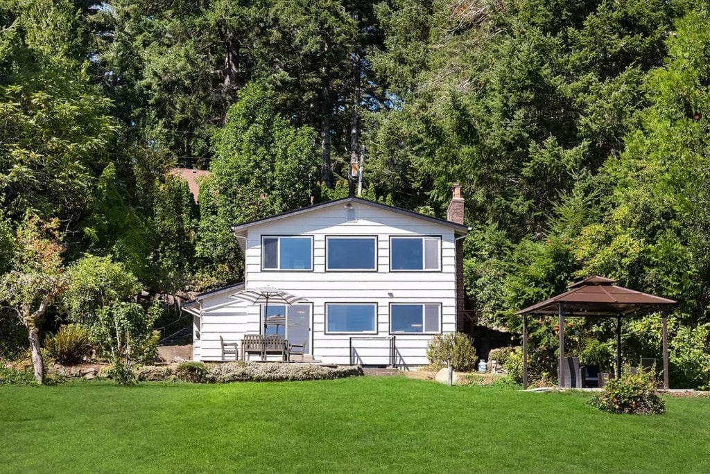 A two-story waterfront home surrounded by dense forest, featuring a large green lawn and an outdoor gazebo, showcasing a diverse property managed in Washington.