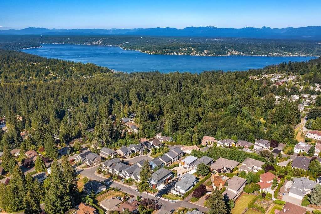 An aerial view of a residential neighborhood surrounded by dense forests and a large body of water, highlighting the scenic location of the developments.