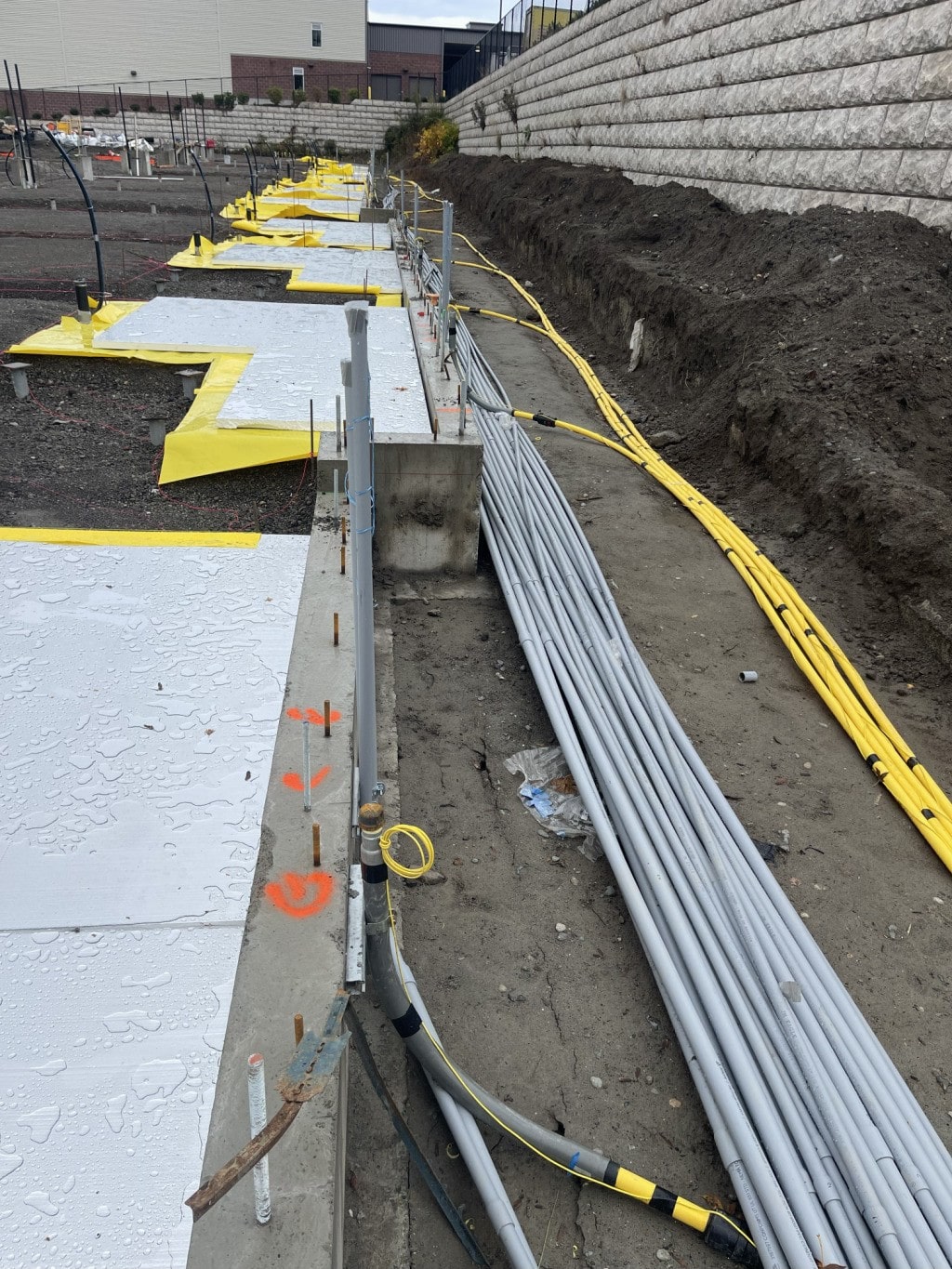 A construction site showing underground utility lines and yellow electrical conduit laid out against a retaining wall, demonstrating infrastructure work.