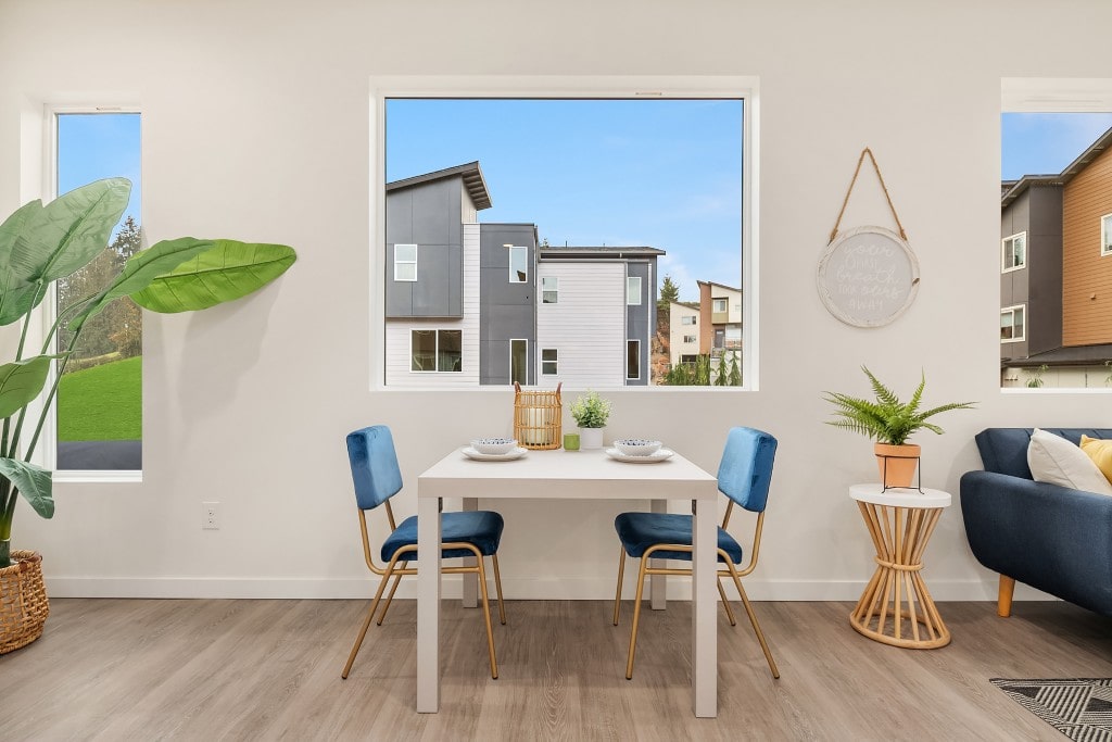 A cozy dining nook with a white table and blue chairs, located next to a large window with a view of the modern townhouse community.