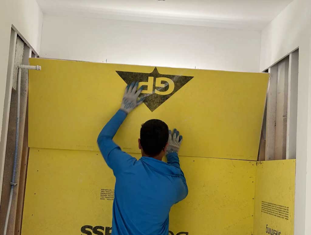 A worker installs yellow drywall or sheathing, showing an interior framing and finishing stage of a new townhouse.