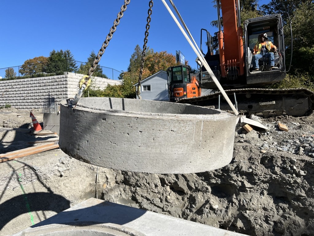 A heavy excavator lifts a large concrete manhole ring into place, showcasing specialized equipment used for utility installation by excavation partners.