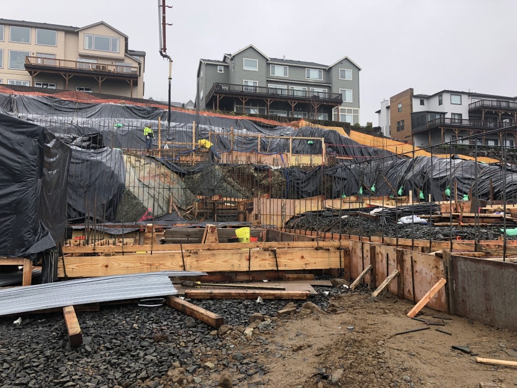 Construction site with workers on a tarp-covered slope, surrounded by wooden frames and gravel. Houses visible above; overcast sky.
