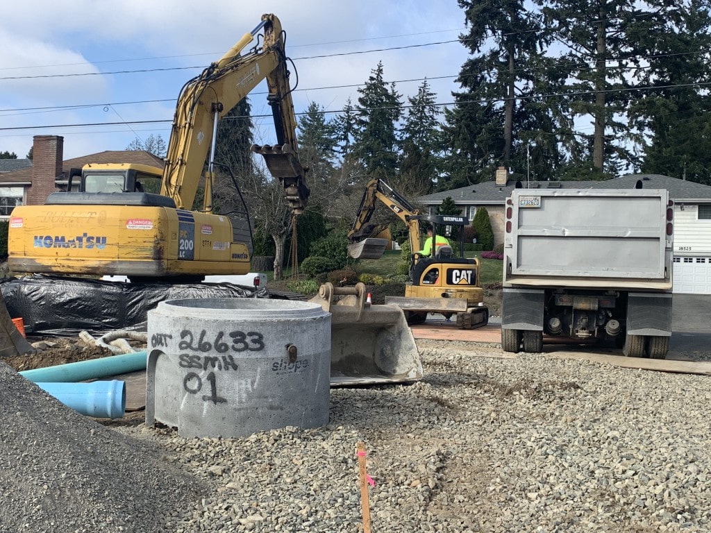 Heavy machinery, including a Komatsu excavator, at a site with a concrete manhole and utility pipes, demonstrating coordinated excavation work.