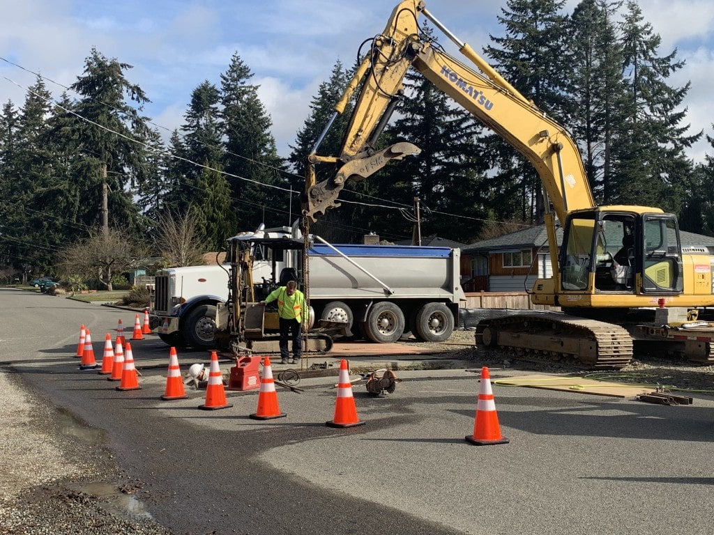 A Komatsu excavator with an attachment works near a dump truck on a street, highlighting the specialized tools used for excavation and material handling.