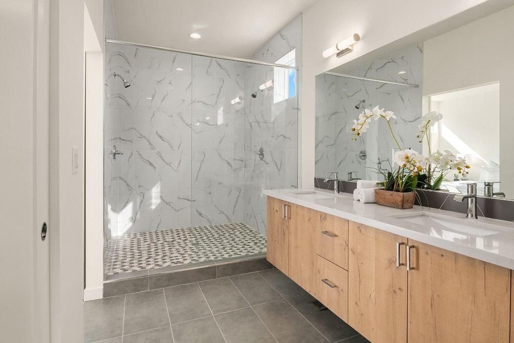 A spacious master bathroom with a double vanity, a large walk-in glass shower with marble-look tile, and modern fixtures, built by ADHG.