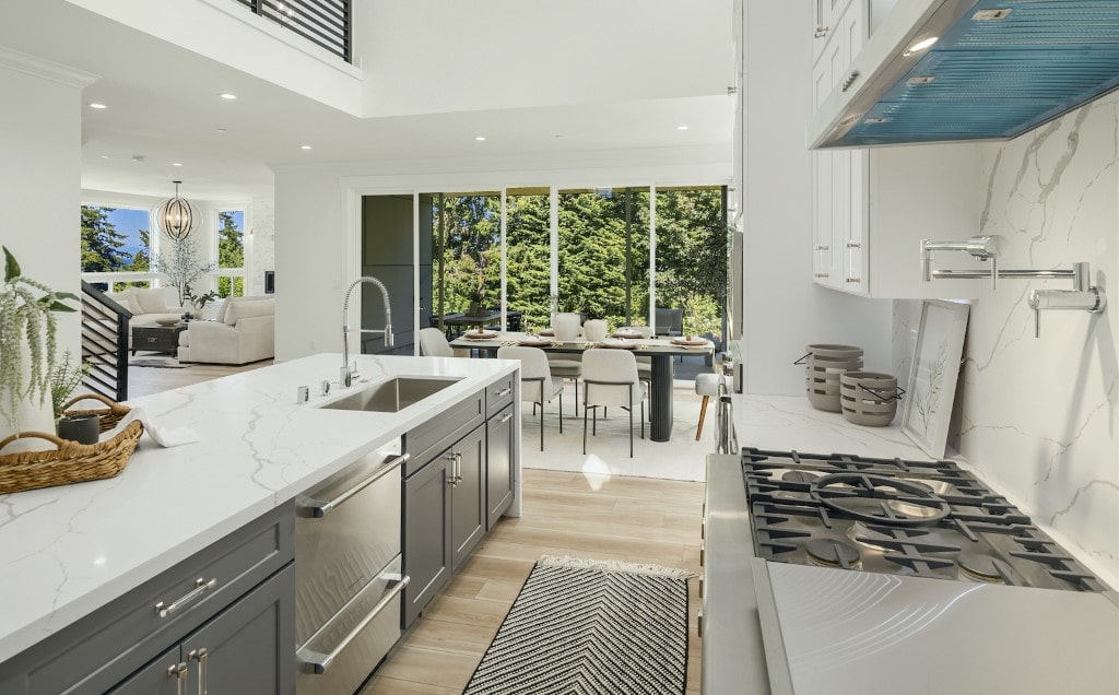 An open-concept kitchen with gray cabinets and white countertops, offering a view into a spacious dining area with large windows, showcasing a functional layout.